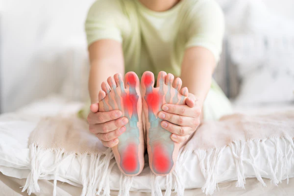 A woman sits in bed, touching her feet, and her feet indicate blue and red spots.